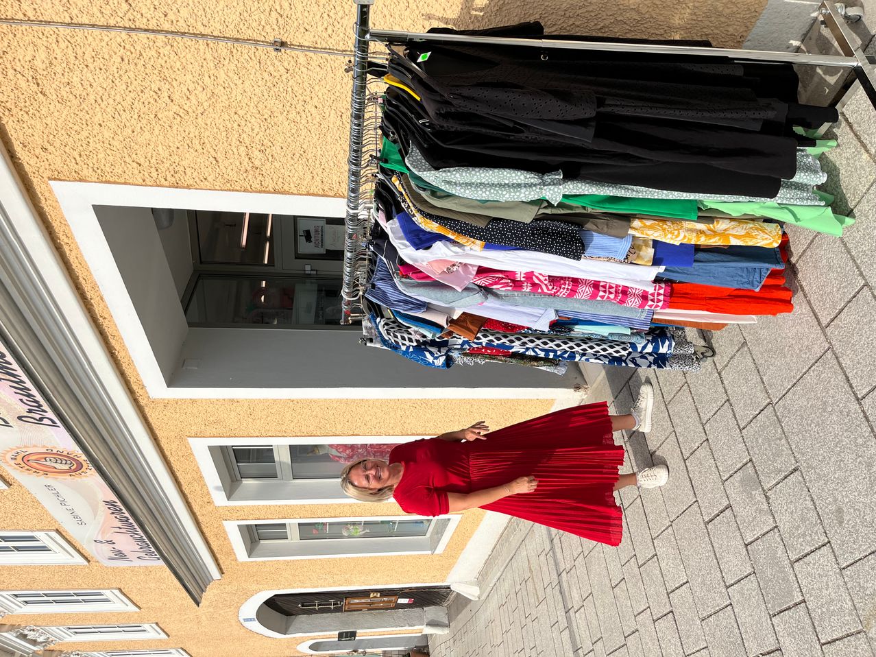 Eine Frau steht vor einem Gebäude mit einem Kleiderständer. Sie trägt ein rotes Kleid und weiße Schuhe.