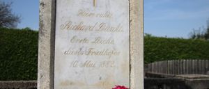 Ein Grabstein mit einem Kreuz und goldener Schrift, datiert auf den 10. Mai 1882, vor einem Busch.