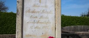 Ein Marmorgrabstein mit einem Kreuz oben, beschriftet mit dem Namen Richard Dunkel, dem Todesdatum 10. Mai 1882 und der Phrase 'Hier ruht'. Eine rote Blume ist vor dem Grabstein platziert.