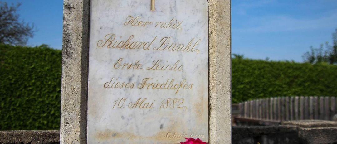Ein Marmorgrabstein mit einem Kreuz oben, beschriftet mit dem Namen Richard Dunkel, dem Todesdatum 10. Mai 1882 und der Phrase 'Hier ruht'. Eine rote Blume ist vor dem Grabstein platziert.