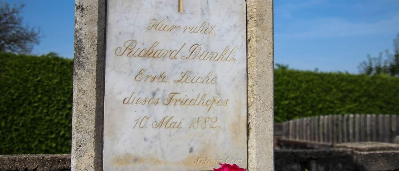Ein Marmorgrabstein mit einem Kreuz oben, beschriftet mit dem Namen Richard Dunkel, dem Todesdatum 10. Mai 1882 und der Phrase 'Hier ruht'. Eine rote Blume ist vor dem Grabstein platziert.