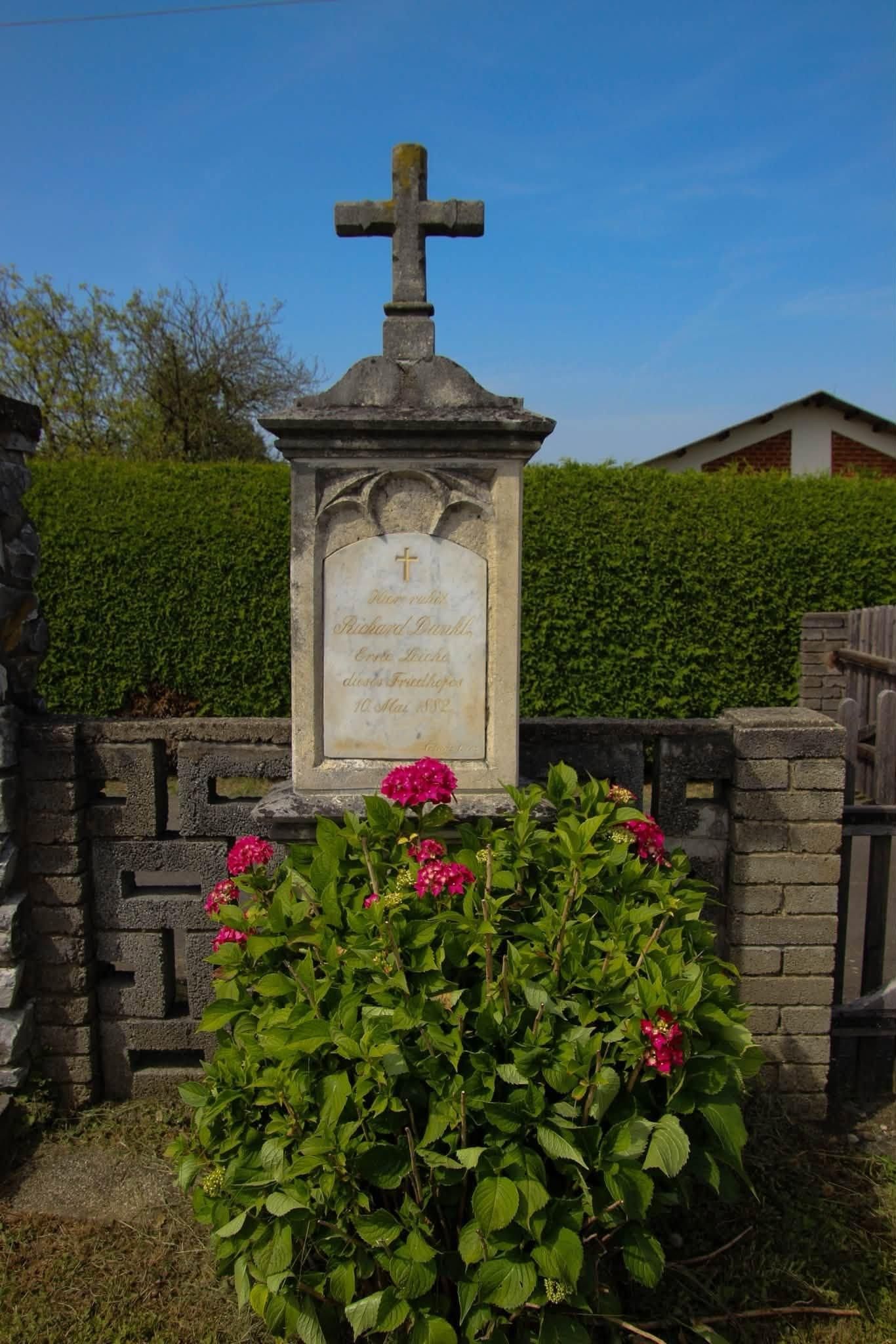 Ein Steinmonument mit einem Kreuz, umgeben von einem Ziegelzaun und Grünpflanzen, mit einer Tafel, die 'Richard Panchl, geboren Lucke, gestorben Fruithof, 10. Mai 1872' besagt.