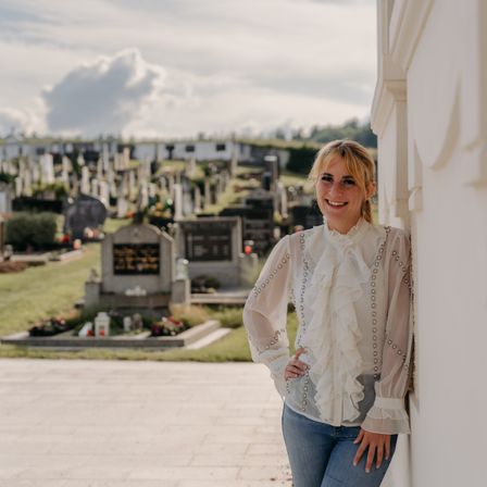 Eine Frau steht in einem Friedhof und lächelt in die Kamera. Sie lehnt sich mit den Händen auf der Hüfte an eine Wand.