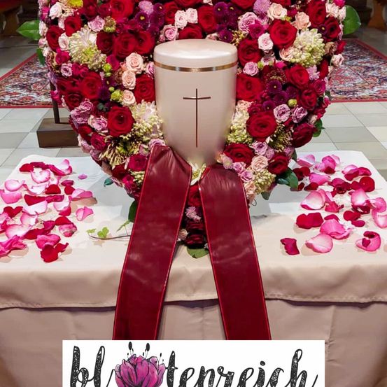 Ein herzförmiges Blumenarrangement mit roten, weißen und rosa Blumen umgibt eine Kreuz-gekrönte Urne, verziert mit verstreuten Rosenblättern auf einem abgedeckten Tisch.