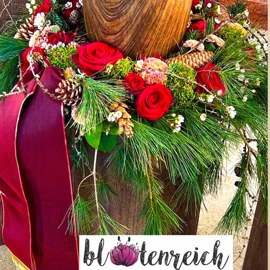 Ein hölzerner Pfosten ist mit einem Kranz aus roten Rosen, Tannenzapfen und Grün geschmückt. Der Kranz besteht aus verschiedenen saisonalen Pflanzen. Der Pfosten ist braun und hat ein Wasserzeichen mit dem Text 'blottenreich'.