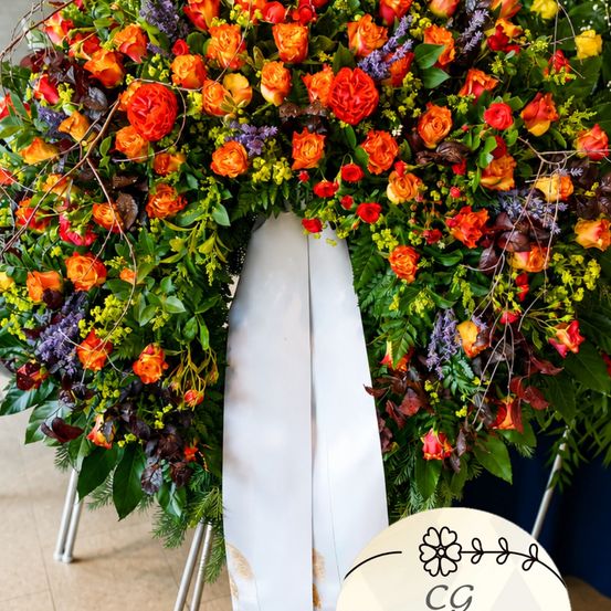 Ein großer Kranz mit leuchtenden roten, orangefarbenen und gelben Blumen, verziert mit grünen Blättern und einer weißen Schleife, auf einem weißen Ständer ausgestellt.