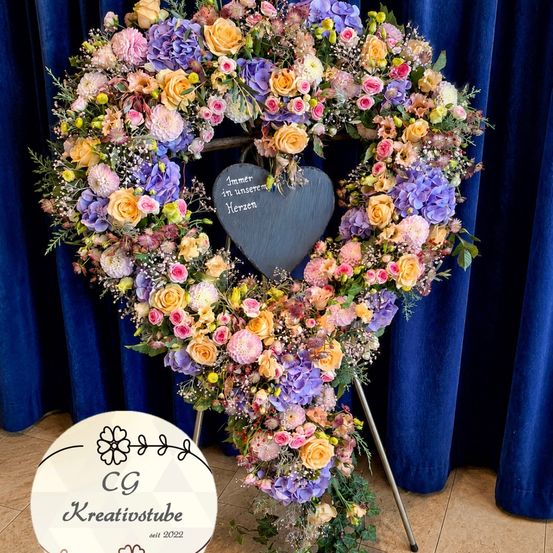 Ein herzförmiger Blumenkranz mit einem Herz in der Mitte steht vor blauen Vorhängen. Auf dem Herz steht 'Immer in unserem Herzen'. Darunter befindet sich ein weißer runder Schild, der 'CG Kreativstübchen seit 2022' besagt.