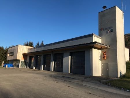 Ein modernes Feuerwehrhaus mit einem hohen Turm und mehreren Garagen, vor dem Hintergrund von Bäumen und einem klaren blauen Himmel.