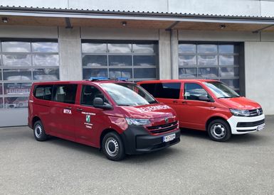 Zwei rote Volkswagen T6-Transporter mit Notfalldienstaufschriften sind vor einem Gebäude mit großen Fenstern geparkt.