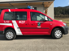 Ein rotes und weißes Notfallfahrzeug mit 'MZF' und einem Logo an der Seite, geparkt vor einem Gebäude mit einer Statue und Bergen im Hintergrund.