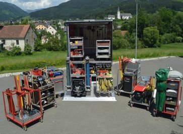 Ein LKW mit geöffnetem Heck, auf dem verschiedene Werkzeuge und Geräte auf Regalen und Trolleys ausgestellt sind. Im Hintergrund ist ein kleines Dorf mit Häusern und einer Kirche zu sehen.