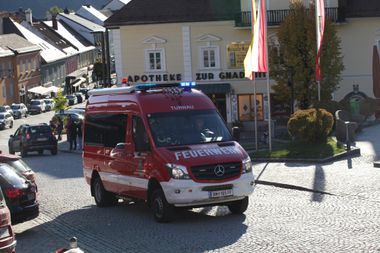 Ein roter und weißer Mercedes-Benz-Van mit dem Wort 'Feuerwehr' auf der Seite fährt auf einer Kopfsteinpflasterstraße. Im Hintergrund befindet sich ein Gebäude mit einer Apotheken-Schild.