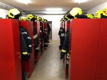 Eine Reihe roter Schließfächer mit ordentlich organisierten Feuerwehruniformen und gelben Helmen. Die Schließfächer sind geschlossen, und die Helme sind oben platziert. Der Raum ist gut beleuchtet und sauber.