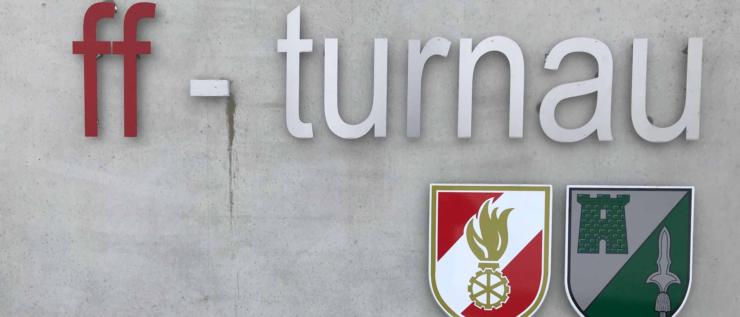 Eine Betonwand zeigt das Wort 'turnau' in weißen Buchstaben mit einem roten 'f'. Unten befinden sich zwei Logos: eines mit einer Flamme und einem Zahnrad, das andere mit einem Burgdesign.