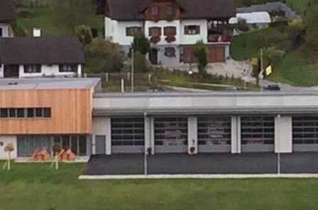 Ein Luftbild einer modernen Feuerwehrstation mit großen Garagentoren und einer Holzfassade. Umgeben sind mehrere Wohnhäuser mit Bäumen und Grünflächen.
