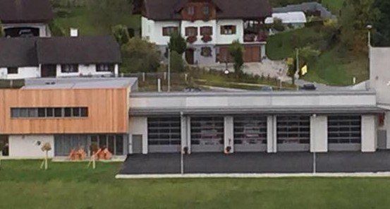 Ein Luftbild einer modernen Feuerwehrstation mit großen Garagentoren und einer Holzfassade. Umgeben sind mehrere Wohnhäuser mit Bäumen und Grünflächen.
