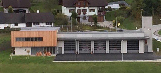 Ein Luftbild einer modernen Feuerwehrstation mit großen Garagentoren und einer Holzfassade. Umgeben sind mehrere Wohnhäuser mit Bäumen und Grünflächen.