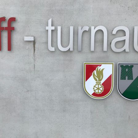 Das Bild zeigt eine Betonwand mit dem Wort 'Turnau' in weißen Buchstaben, geteilt durch ein rotes 'ff'. Darunter befinden sich zwei Logos, eines mit einer Flamme und einem Rad, und ein anderes mit einer Burg.