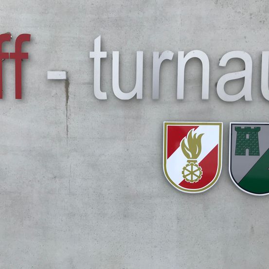 Das Bild zeigt eine Betonwand mit dem Wort 'Turnau' in weißen Buchstaben, geteilt durch ein rotes 'ff'. Darunter befinden sich zwei Logos, eines mit einer Flamme und einem Rad, und ein anderes mit einer Burg.
