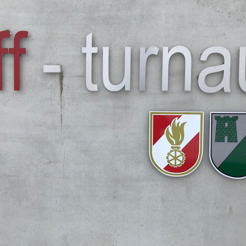 Das Bild zeigt eine Betonwand mit dem Wort 'Turnau' in weißen Buchstaben, geteilt durch ein rotes 'ff'. Darunter befinden sich zwei Logos, eines mit einer Flamme und einem Rad, und ein anderes mit einer Burg.