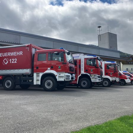 Vier rote Feuerwehrfahrzeuge sind in einer Reihe vor einem Gebäude mit bewölktem Himmel geparkt.