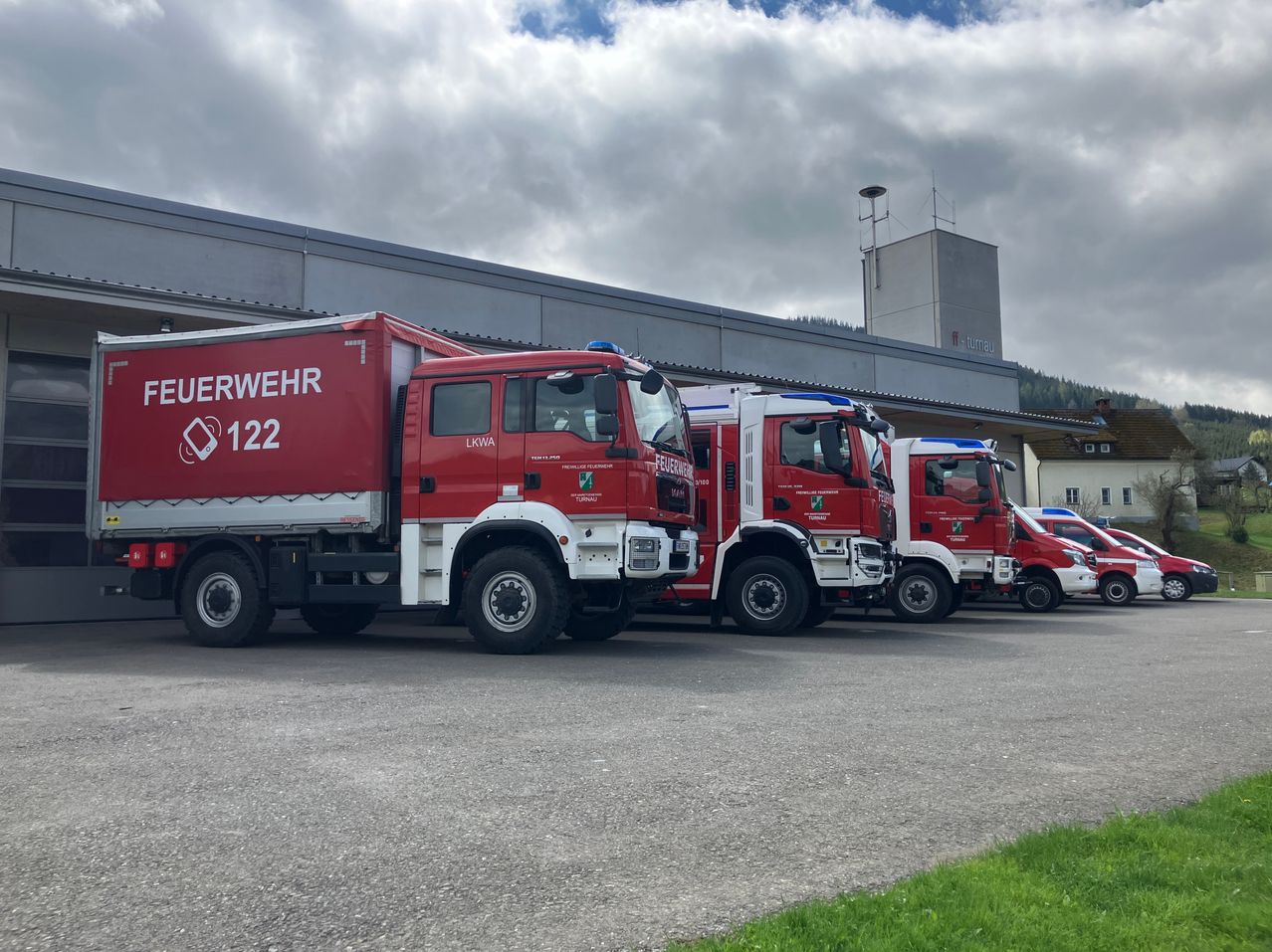 Vier rote Feuerwehrfahrzeuge sind in einer Reihe vor einem Gebäude mit bewölktem Himmel geparkt.