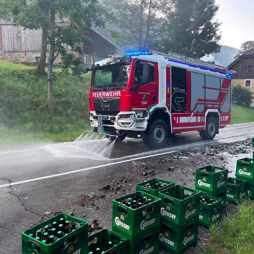 Ein roter Feuerwehrwagen spükte Wasser auf die Straße, umher lagen mehrere Kisten Güsser Bier. Die Szene spielt sich in einem ländlichen Gebiet mit einem grasigen Hang im Hintergrund ab.