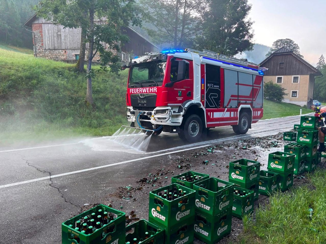 Ein roter Feuerwehrwagen spükte Wasser auf die Straße, umher lagen mehrere Kisten Güsser Bier. Die Szene spielt sich in einem ländlichen Gebiet mit einem grasigen Hang im Hintergrund ab.
