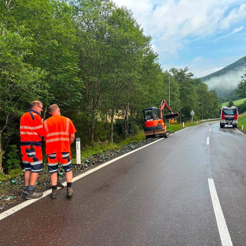 Zwei Männer in orangefarbenen Uniformen stehen am Straßenrand und beobachten ein geparktes Baufahrzeug. Ein LKW fährt auf der Straße. Bäume und Berge sind im Hintergrund.