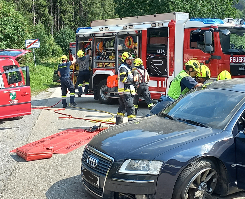 Feuerwehrleute in voller Ausrüstung arbeiten an der Unfallstelle eines Autos, wobei eine Person im Fahrzeug sitzt und andere sich um Ausrüstung auf einem Feuerwehrwagen kümmern.