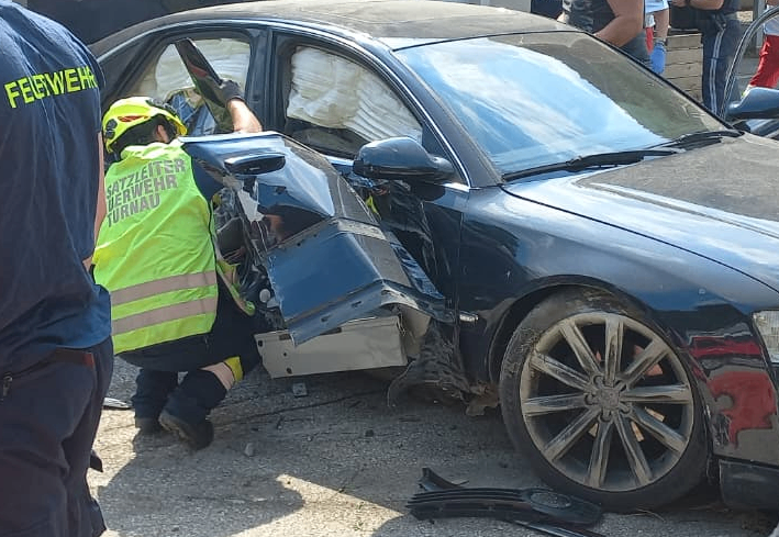 Ein Rettungsarbeiter in einer gelben Weste entfernt eine Person aus einem beschädigten blauen Auto. Das Auto hat eine zertrümmerte Front und ist von Trümmern umgeben.