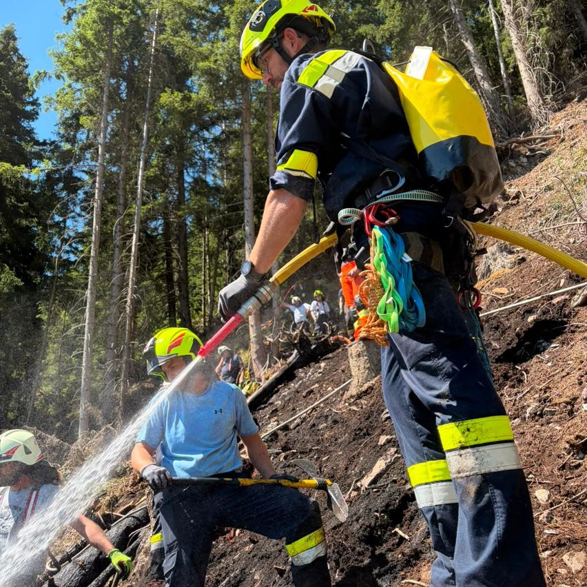 Feuerwehrleute in Schutzkleidung arbeiten an einem Hang, einer spritzt Wasser, während der andere ein Werkzeug hält. Bäume und ein klarer Himmel sind im Hintergrund zu sehen.