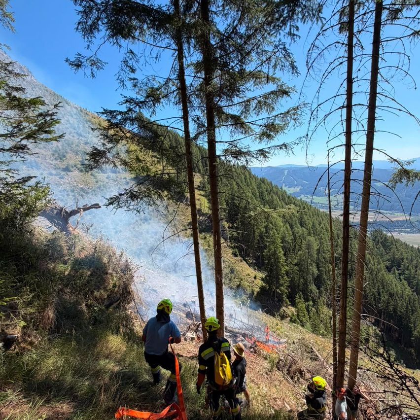 Eine Gruppe von Feuerwehrleuten in Schutzkleidung befindet sich auf einem Hang mit einem brennenden Bereich im Hintergrund.