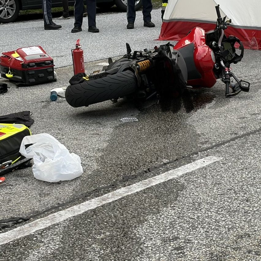 Ein rotes Motorrad liegt auf der Straße, neben einem Feuerlöscher und einem roten Erste-Hilfe-Set. Im Hintergrund steht ein weißes Zelt und ein schwarzes Auto.
