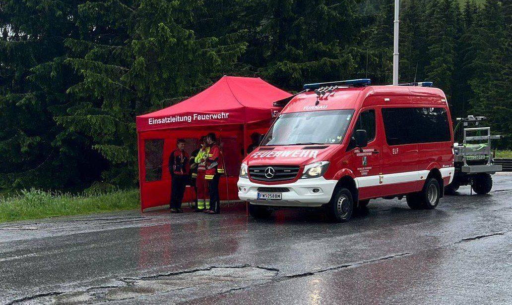 Ein roter Feuerwehrwagen ist neben einem roten Zelt mit der Aufschrift 'Einsatzleitung Feuerwehr' geparkt. Vier Feuerwehrleute sind im Zelt. Die Straße ist nass.