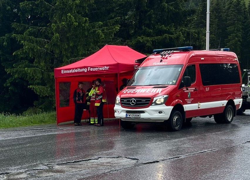 Ein roter Feuerwehrwagen ist neben einem roten Zelt mit der Aufschrift 'Einsatzleitung Feuerwehr' geparkt. Vier Feuerwehrleute sind im Zelt. Die Straße ist nass.