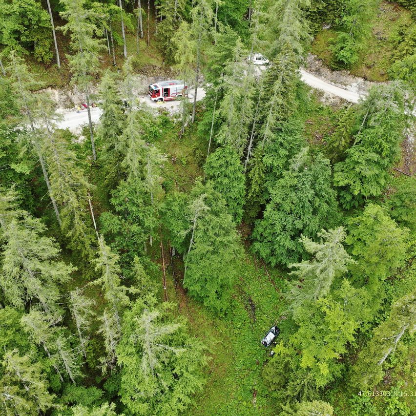 Luftaufnahme eines Unfallortes im Wald. Ein Rettungsfahrzeug ist am Straßenrand geparkt. Ein Auto ist vom Weg abgekommen und im Wald verunglückt.