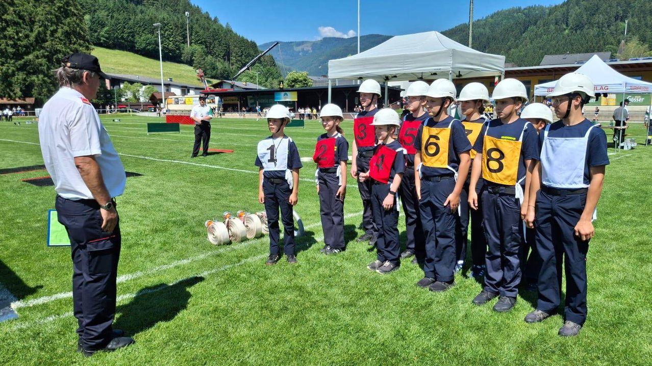 Eine Gruppe von Kindern in gleichen Uniformen und Helmen steht auf einem Feld, nummeriert von 1 bis 8. Eine Person steht hinter ihnen, und ein Zelt ist in der Nähe aufgespannt. Im Hintergrund sind Berge zu sehen.