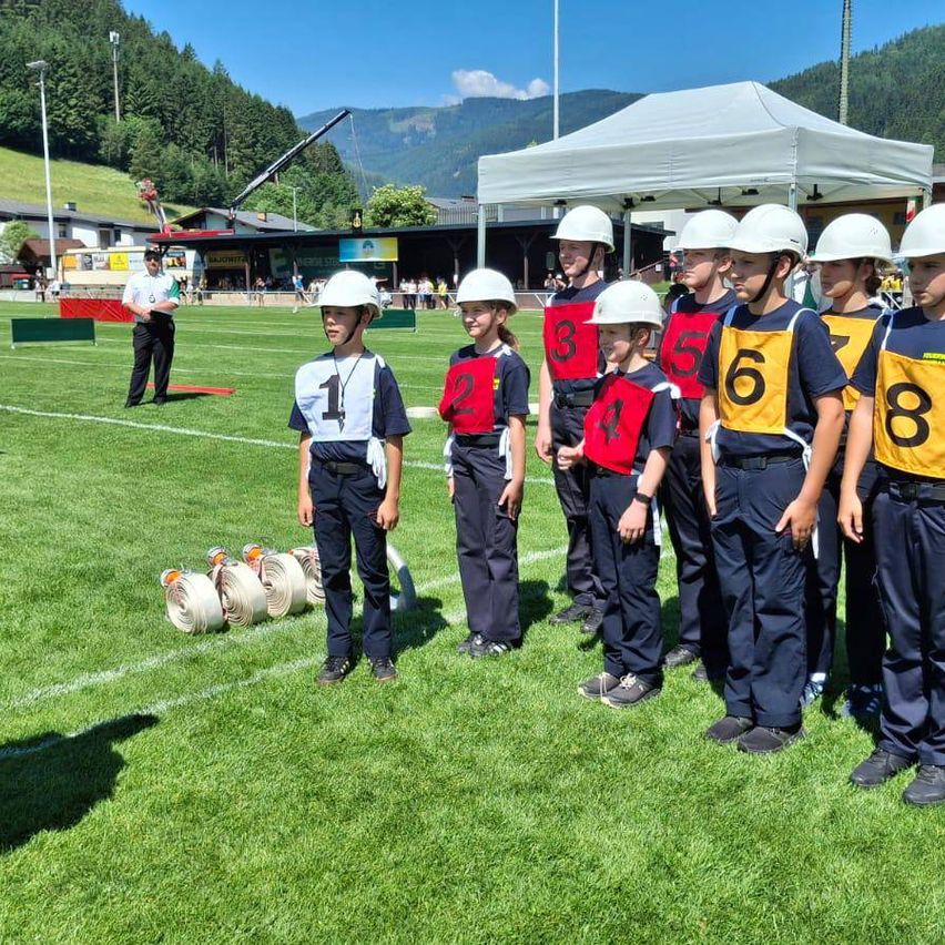Eine Gruppe von Kindern in gleichen Uniformen und Helmen steht auf einem Feld, nummeriert von 1 bis 8. Eine Person steht hinter ihnen, und ein Zelt ist in der Nähe aufgespannt. Im Hintergrund sind Berge zu sehen.