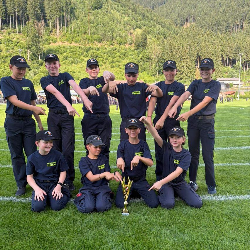 Ein Team junger Athleten, in einheitlichen Trikots gekleidet, posiert mit einem Pokal auf einem Rasenfeld, mit einem bewaldeten Hügel im Hintergrund.