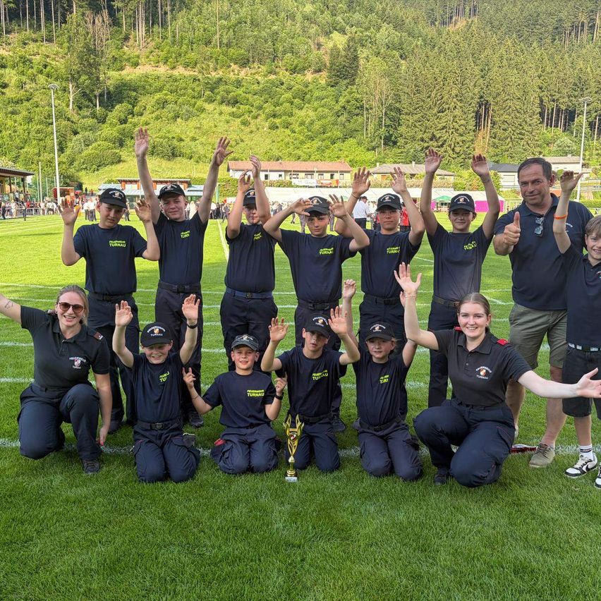 Eine Gruppe Feuerwehrleute in Uniform posiert für ein Foto auf einem Rasenfeld, hält eine Trophäe in der Hand, mit einem bewaldeten Hügel im Hintergrund.