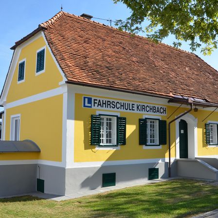 Ein gelbes Gebäude mit braunem Dach namens Fahrschule Kirchbach. Es hat grüne Fensterläden und eine schwarze Tür, mit einem Rasenbereich davor.