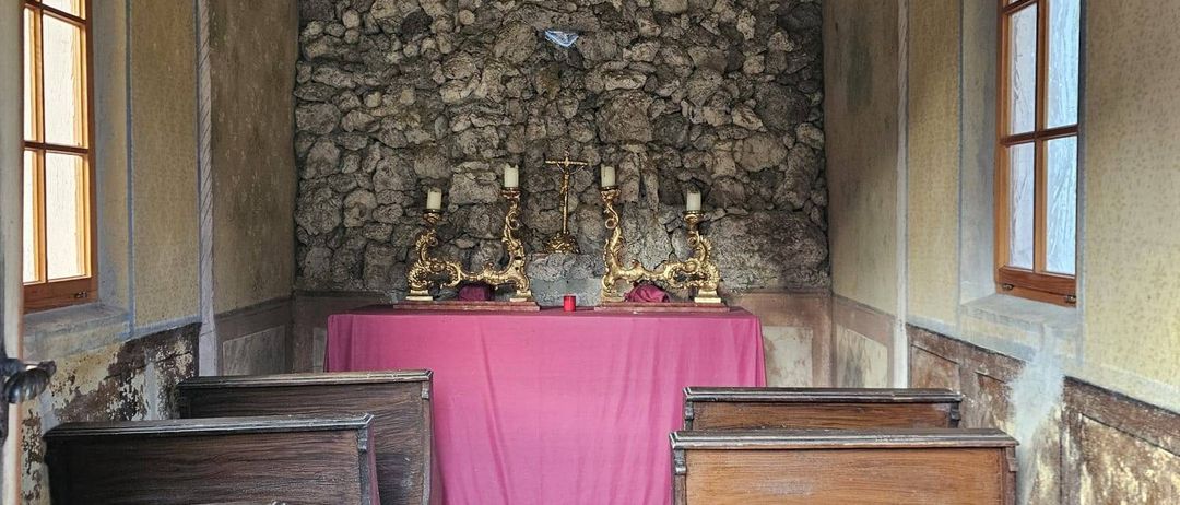 Eine kleine Kapelle mit Holzbänken, die auf einen Tisch mit einem rosa Tuch zeigen. Drei goldene Kerzenhalter stehen auf dem Tisch.