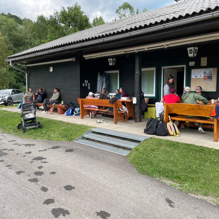 Eine Gruppe von Menschen sitzt auf Bänken vor einem Gebäude mit einer grünen Wand und einem Rasenbereich. Ein Kinderwagen steht links, und ein Auto ist dahinter geparkt. Bäume und Berge sind im Hintergrund.
