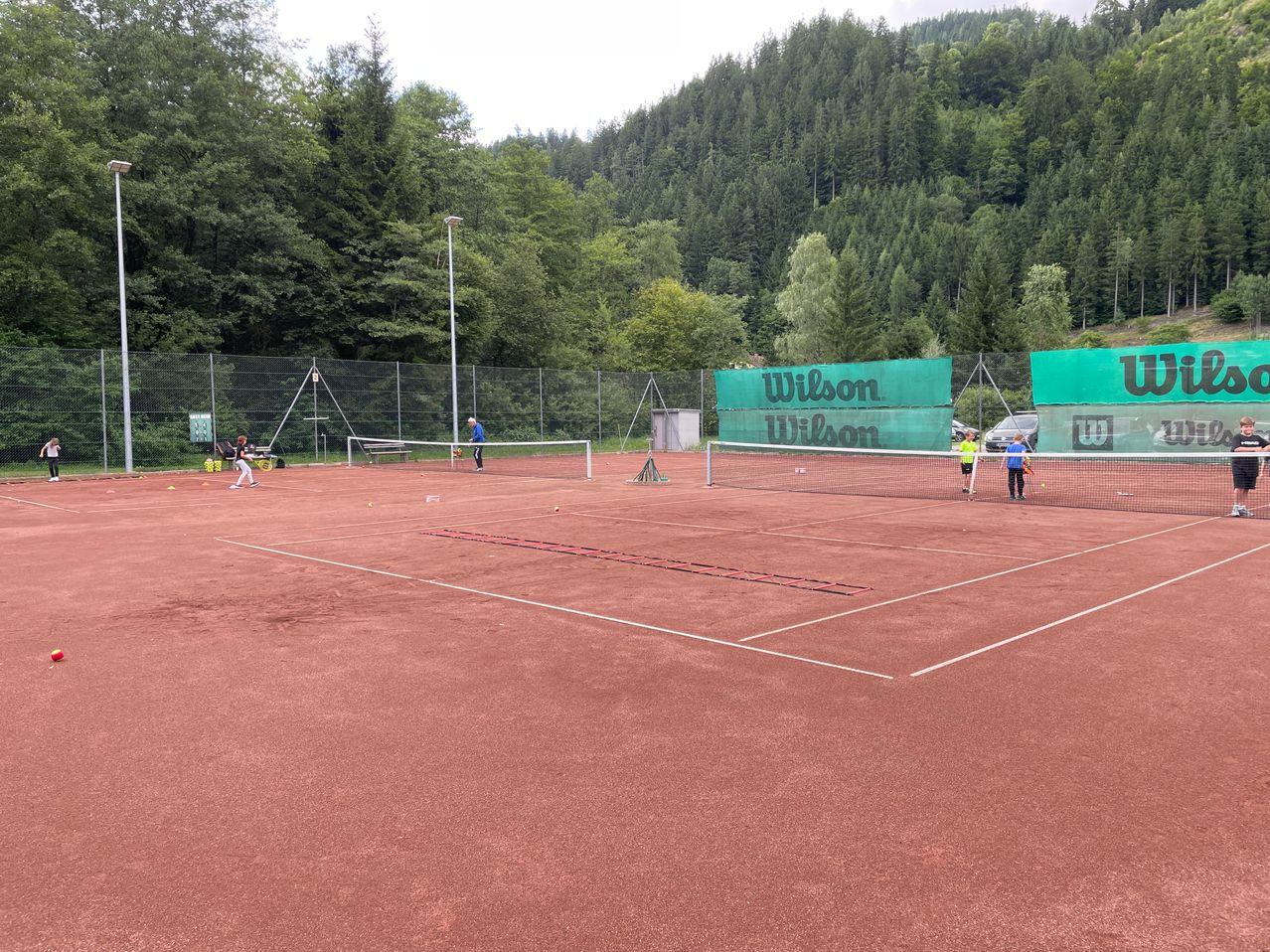 Tennisspieler trainieren auf einem Platz mit rotem Untergrund. Wilson-Werbung befindet sich an den Seitenzäunen.
