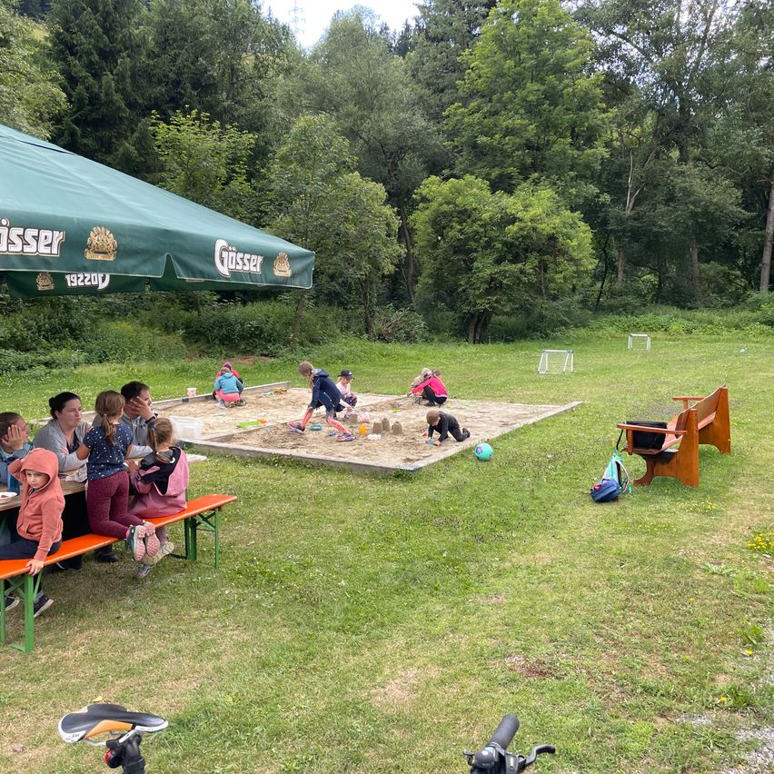 In einem Park spielen eine Gruppe von Kindern in einem Sandkasten unter einem grünen Schirm, während Erwachsene auf Bänken sitzen. Bäume und ein Fußballtor sind im Hintergrund.