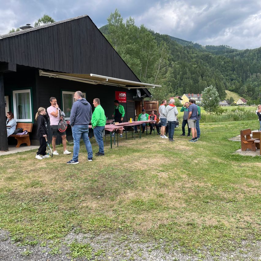 Mehrere Menschen versammeln sich vor einem Gebäude mit einem Schild, Tischen mit Essen und einem Tennisplatz. Im Hintergrund befinden sich Bäume, Berge und Häuser.