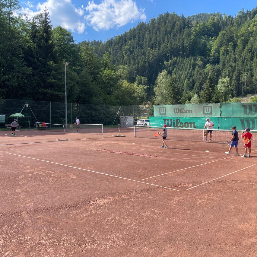 A group of people are playing tennis on a dirt court with a mountain in the background. There are four tennis nets set up, and players are positioned on each side.