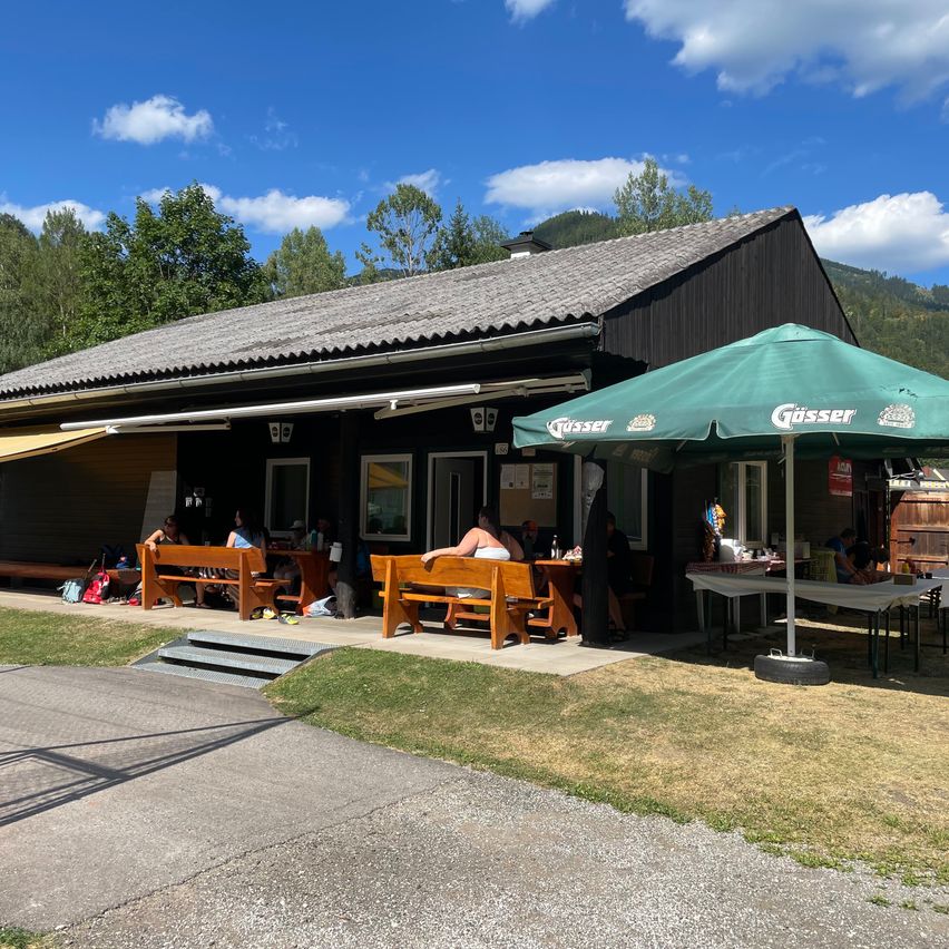 A rustic outdoor dining area with wooden benches and a green umbrella in front of a black building with a gray roof. Several people are seated at tables under the umbrella.