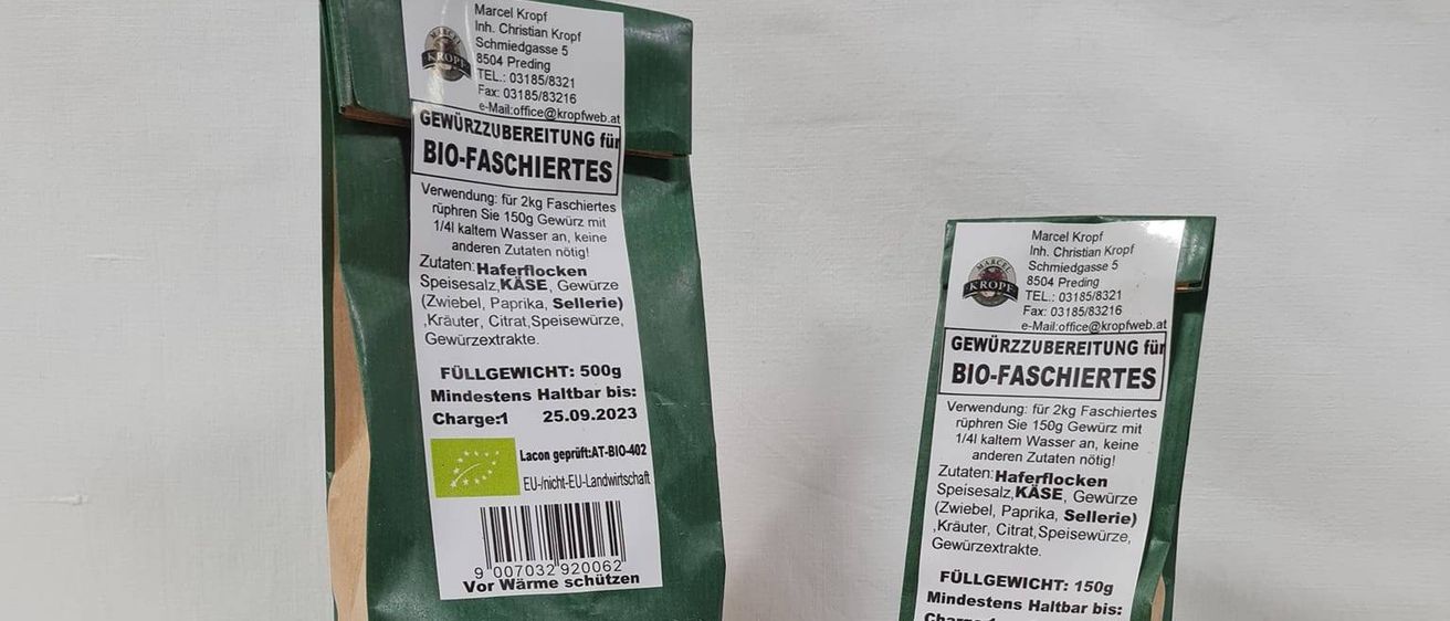 Zwei Beutel Bio-Gewürzmischung, einer geöffnet und einer geschlossen, liegen auf einem Tisch. Daneben befinden sich ein Teller mit Gewürzmischung und ein Teller mit geschnittenem Fleisch.